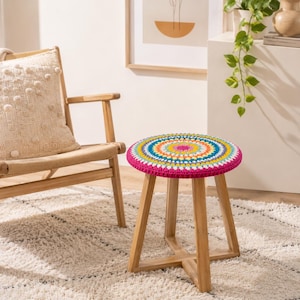 May include: A wooden stool with a round, colorful crocheted seat. The seat features a vibrant pattern of concentric circles in shades of pink, yellow, blue, and green. The stool has three wooden legs and is set on a cream-colored rug.