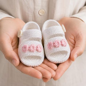 Op de afbeelding: Een paar witte gehaakte babysandalen met roze bloemaccenten. De sandalen hebben een knoopsluiting en worden in open handen gehouden. Een handgemaakt artikel.