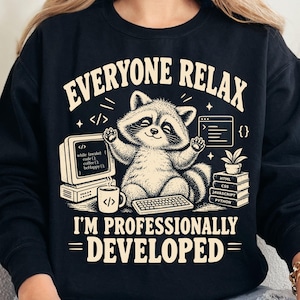 Może przedstawiać: Czarna bluza z długim rękawem z grafiką szopa pracza siedzącego przy komputerze oraz napisem "EVERYONE RELAX I'M PROFESSIONALLY DEVELOPED". Wzór zawiera monitor z kodem, klawiaturę, kubek z kawą oraz stos książek z nazwami języków programowania.