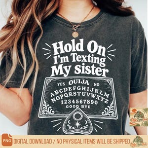 May include: A charcoal gray t-shirt featuring white screen-printed text that reads Hold On I'm Texting My Sister above a graphic of a Ouija board with the alphabet, numbers zero through nine, and a planchette centered at the bottom of the design.