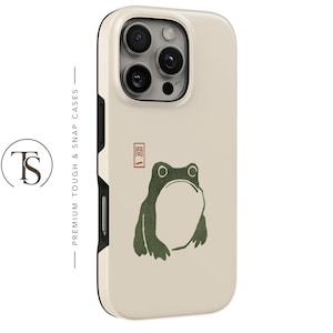 May include: A cream-colored phone case featuring a dark green frog illustration and a small red rectangular stamp with Japanese characters. The case has a black inner frame and a triple-lens camera cutout. The text "PREMIUM TOUGH & SNAP CASES" is visible.
