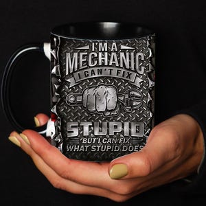 May include: Black ceramic mug with a black handle and rim. The mug features the text "I'M A MECHANIC I CAN'T FIX STUPID BUT I CAN FIX WHAT STUPID DOES" in a silver, industrial-style design with a wrench and fist graphic.