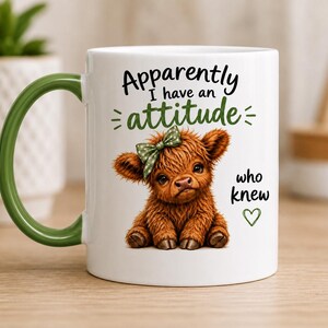 May include: A white ceramic mug with a green handle featuring a graphic of a fluffy brown Highland calf wearing a green polka-dot bow. The text reads "Apparently I have an attitude who knew" in black and green, accompanied by a small green heart icon.