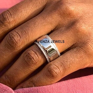 Puede incluir: Un anillo ancho de color plateado con un centro rectangular y una fila de pequeñas piedras transparentes a cada lado. El anillo está en una mano, y el texto "AVENZA JEWELS" es visible.
