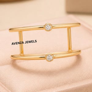 Könnte beinhalten: Ein goldfarbener Ring mit einem einzigartigen Doppelband-Design. Der Ring hat zwei kleine, runde, klare Edelsteine, die in das Band eingesetzt sind. Der Text "AVENZA JEWELS" ist auf dem Ring aufgedruckt.