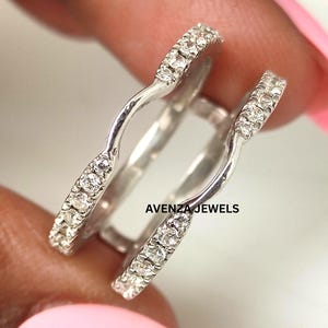 May include: Two silver rings with a curved design, each encrusted with small, clear gemstones. The rings are held up, showcasing their shape and sparkle. The text "AVENZA JEWELS" is visible.