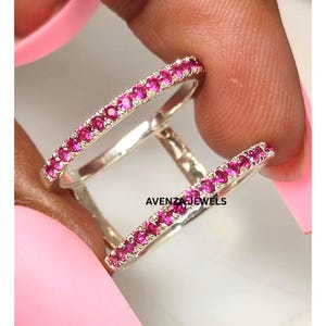 Puede incluir: Dos anillos de plata con una fila de pequeñas piedras preciosas de color rosa brillante. Los anillos se sostienen entre los dedos, con el nombre de la marca "AVENZA JEWELS" visible.