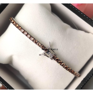 Pode incluir: Uma pulseira delicada com uma fileira de pedras preciosas castanhas e claras. A pulseira é definida em metal prateado e é exibida em uma caixa preta com uma almofada branca. O texto "AVANZA JEWELS" é visível.