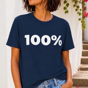 Op de afbeelding: Marineblauw T-shirt met de tekst "100%" in het wit. Het shirt wordt gedragen met lichtblauwe denim jeans. De persoon staat buiten in de buurt van een witte muur en trappen.