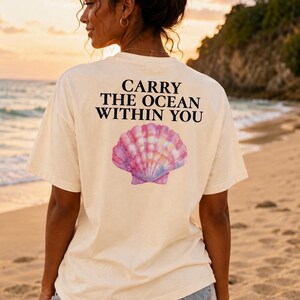 Puede incluir: Una camiseta de manga corta color blanco roto con el texto en negro CARRY THE OCEAN WITHIN YOU impreso sobre una ilustración estilo acuarela de una concha marina rosa, azul y morada en la parte posterior de la prenda.