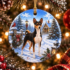 May include: A round ceramic Christmas ornament featuring a tricolor Basenji dog standing in a snowy winter landscape. The scene includes a wooden fence with a wreath, a small cabin, pine trees, and a lantern, all suspended by a gold cord.
