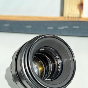May include: A black camera lens with the text "Гелиос-44-2" printed on the side. The lens has a ridged focus ring and a clear glass element. A black lens cap is visible in the foreground.