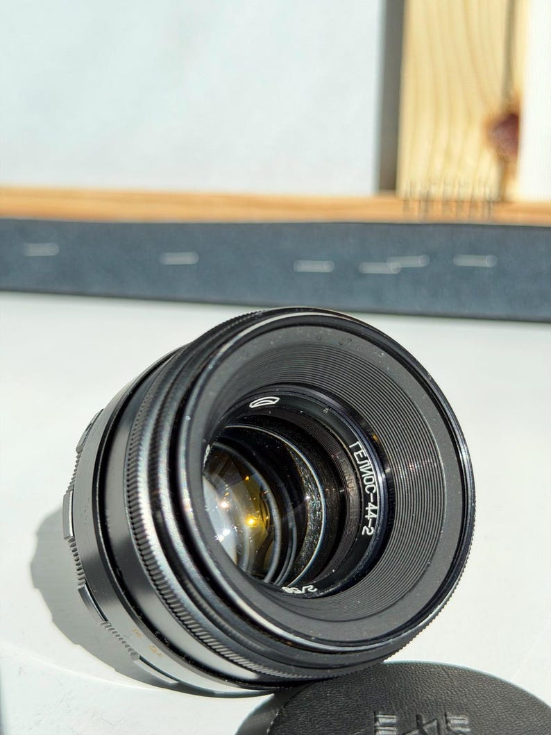 May include: A black camera lens with the text "Гелиос-44-2" printed on the side. The lens has a ridged focus ring and a clear glass element. A black lens cap is visible in the foreground.