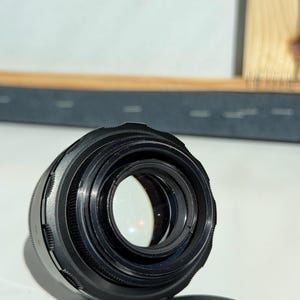 May include: A black camera lens with multiple rings and a clear glass element. The lens is positioned on a white surface, with a black lens cap visible in the foreground. The lens cap has text on it.