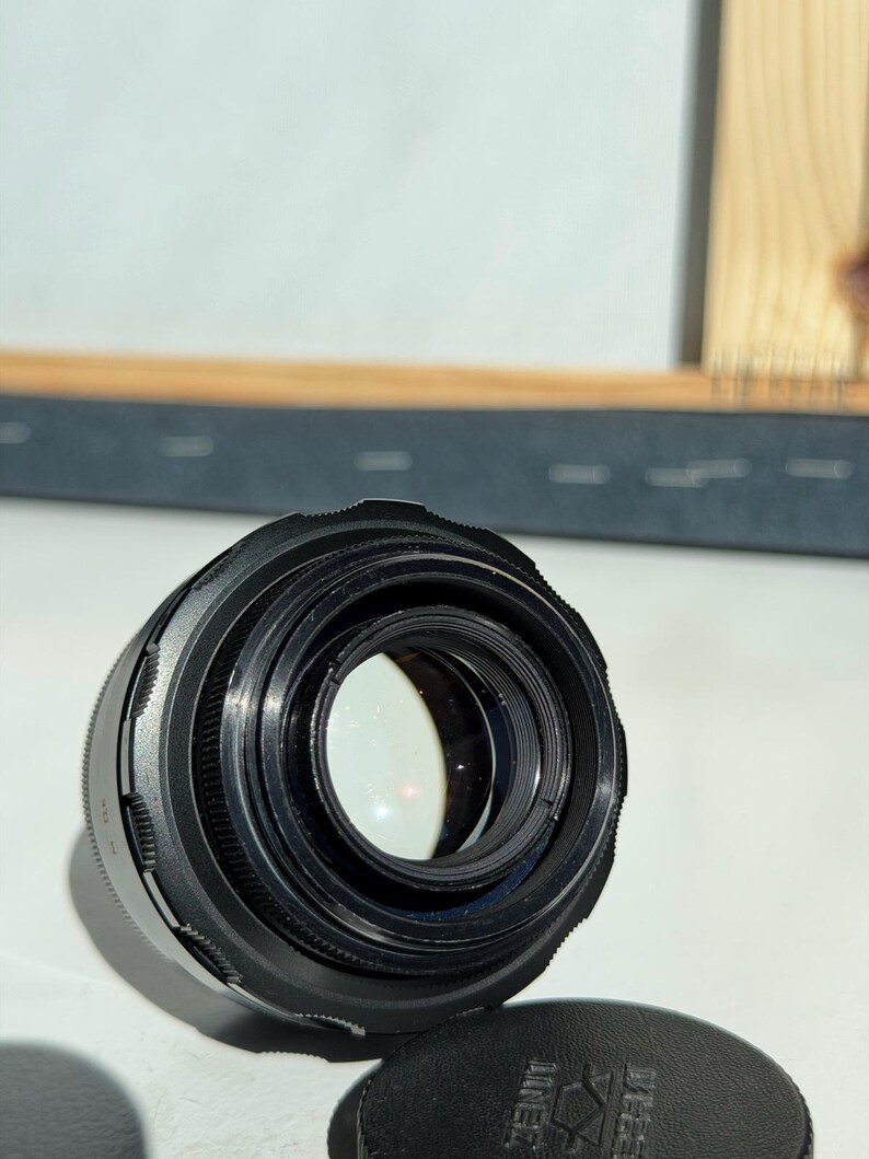 May include: A black camera lens with multiple rings and a clear glass element. The lens is positioned on a white surface, with a black lens cap visible in the foreground. The lens cap has text on it.