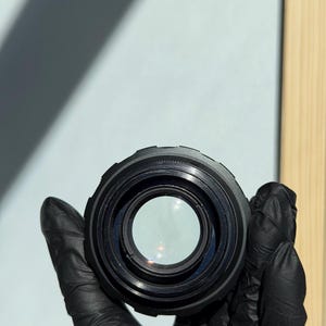 May include: A black camera lens held by a gloved hand. The lens has a circular design with a clear glass center. The lens is a photography accessory.