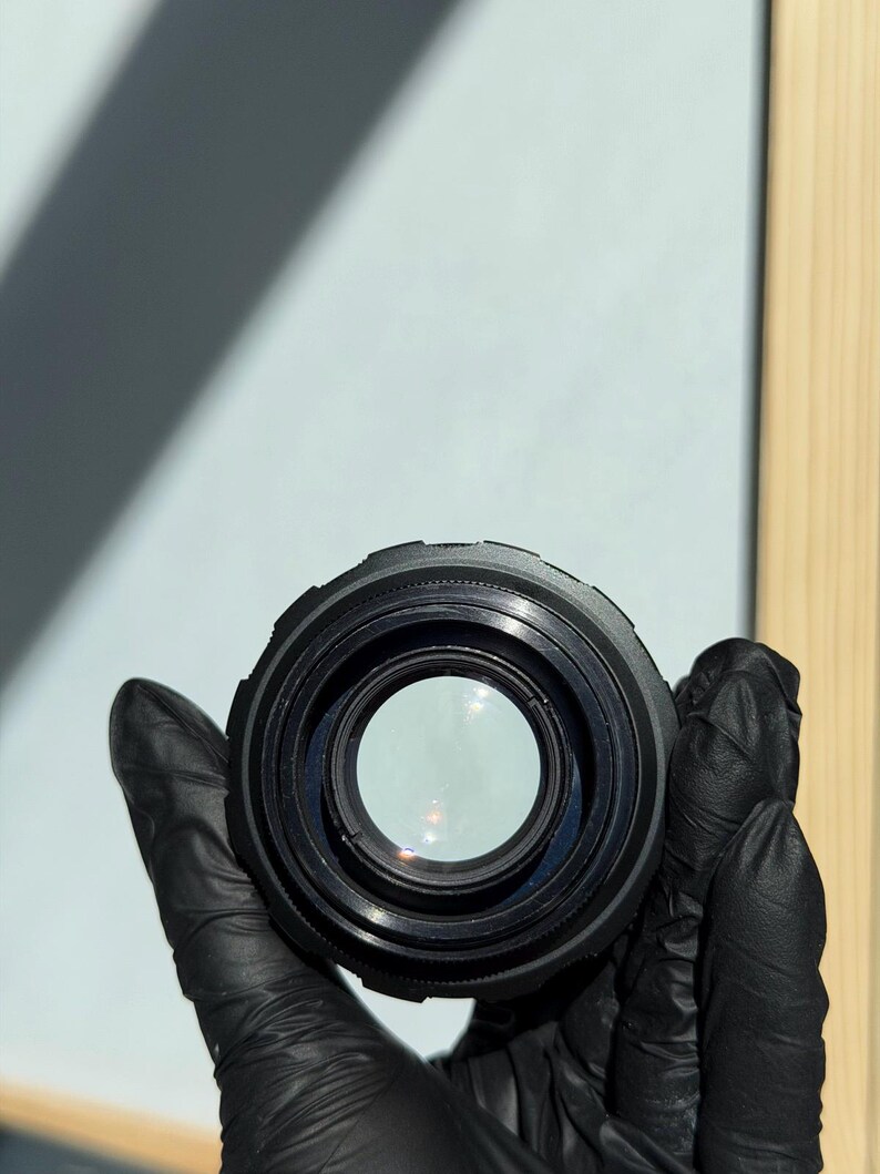 May include: A black camera lens held by a gloved hand. The lens has a circular design with a clear glass center. The lens is a photography accessory.