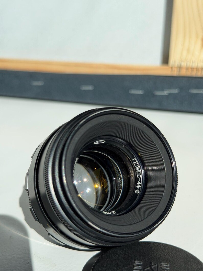 May include: A black camera lens with the text "Гелиос-44-2" on the side. The lens has a ridged focus ring and a reflective interior. A black lens cap is at the bottom.