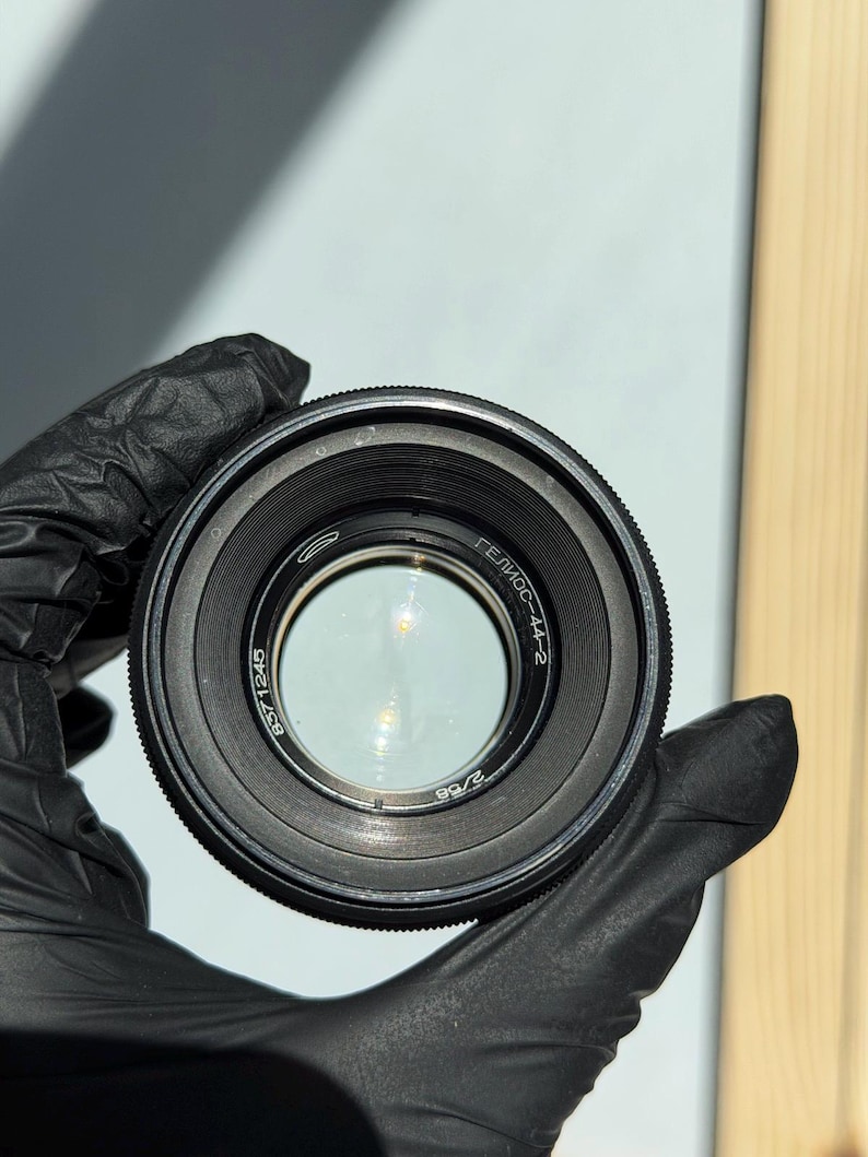 May include: A black camera lens with the text "ГЕЛИОС-44-2" and other markings. The lens has a glass element in the center and is held by a gloved hand. The lens is a photography accessory.