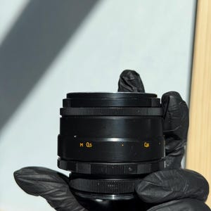May include: A black camera lens with the markings "M 0,5" and "0,6" in yellow. The lens has a ridged design and is held by a gloved hand. The background is a light blue and white.