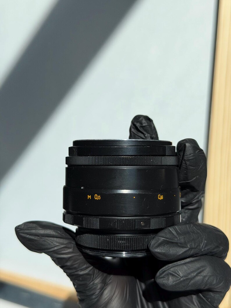 May include: A black camera lens with the markings "M 0,5" and "0,6" in yellow. The lens has a ridged design and is held by a gloved hand. The background is a light blue and white.