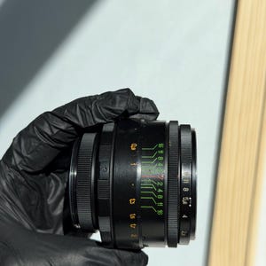 May include: A black camera lens with a series of adjustable rings and markings. The lens has green and white numerical indicators, including f-stop values. The lens is held by a gloved hand against a neutral background.