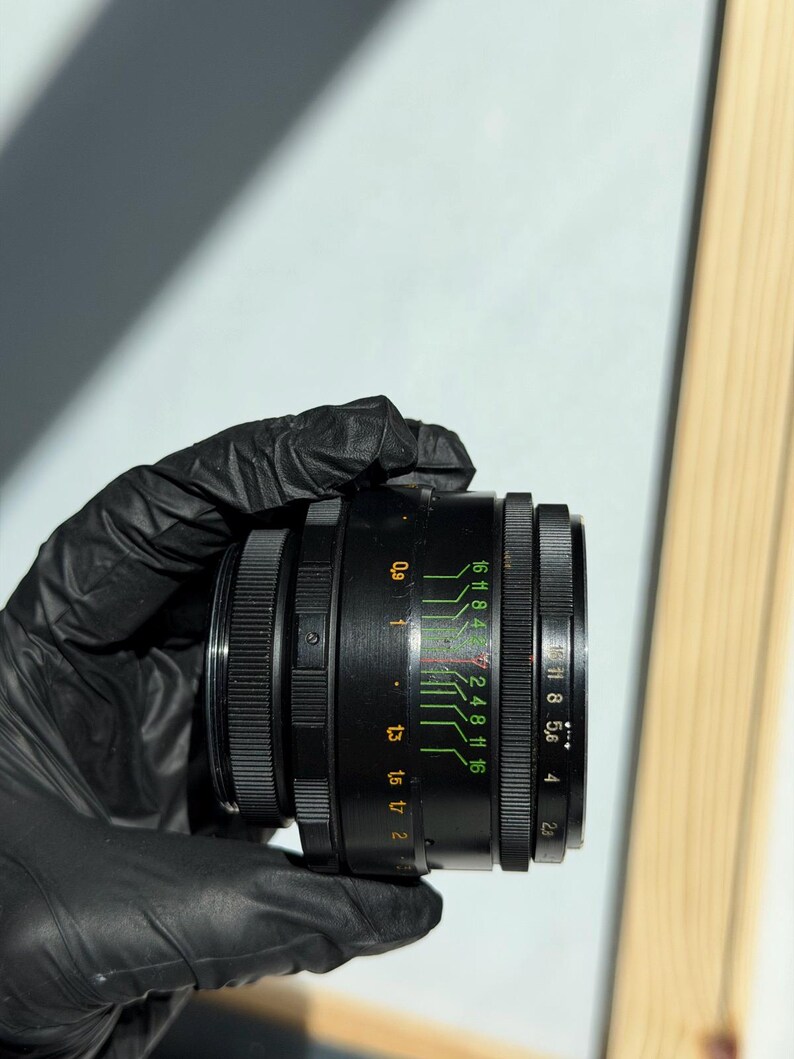 May include: A black camera lens with a series of adjustable rings and markings. The lens has green and white numerical indicators, including f-stop values. The lens is held by a gloved hand against a neutral background.