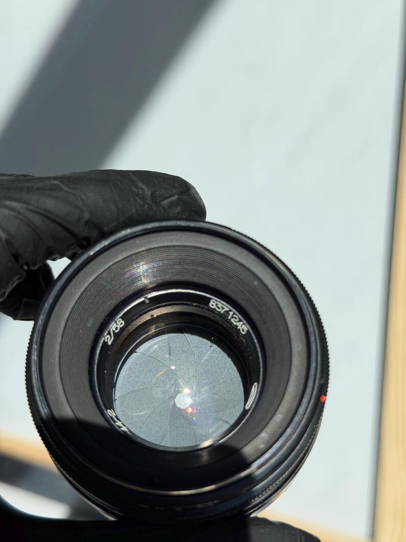 May include: A black camera lens with the numbers "2/58" and "837 1245" visible. The lens has a series of concentric rings and a red dot. The aperture blades are partially closed, creating a star-like pattern.