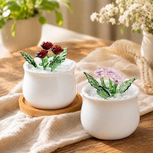 May include: Two small white ceramic pots each hold glass flowers. One pot has red roses, the other has pink flowers. The flowers have green glass leaves and sit in white pebbles. One pot rests on a wooden coaster.