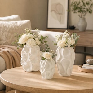 Può includere: Tre vasi in ceramica bianca di diverse dimensioni, ciascuno con fiori bianchi e verde. I vasi hanno superfici strutturate e sono disposti su un tavolo rotondo in legno. Lo sfondo comprende un divano e un'opera d'arte incorniciata.