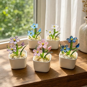 May include: Five small, square white ceramic pots each hold a glass flower arrangement. The flowers are in shades of blue, pink, and purple, with green stems and leaves. The pots sit on a wooden surface near a window.