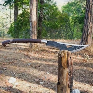 May include: A curved blade knife with a dark brown leather-wrapped handle and a metallic bolster. The blade features a triangular cutout near the spine and is engraved with the text Eagle Cutlery Pakistan. The knife is balanced on a wooden post.