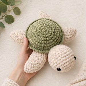 May include: A handmade crochet turtle toy. The turtle has a green shell, cream-colored flippers, and a cream-colored head with black eyes. The toy is made of soft yarn and is designed to be a cuddly item.