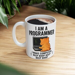 May include: A white ceramic mug featuring a black graphic of an orange tabby cat sitting at a laptop. The text reads I AM A PROGRAMMER and I MAKE COMPUTER BEEP BOOP BEEP BEEP BOOP. The mug is filled with a dark liquid, likely coffee, with visible foam.
