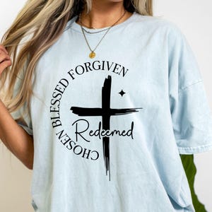 Op de afbeelding: Lichtblauw T-shirt met een zwart kruisontwerp en de woorden "Blessed, Forgiven, Chosen, Redeemed". Het ontwerp is gecentreerd. Het shirt heeft korte mouwen en een ronde hals.