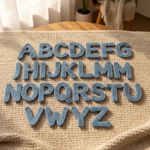 Op de afbeelding: Een volledig alfabet van blauwe gehaakte letters, gerangschikt op een beige, getextureerd oppervlak. De letters zijn in een schreefloze lettertype en lijken handgemaakt. De letters zijn gelijkmatig verdeeld en hebben een zachte blauwe kleur.
