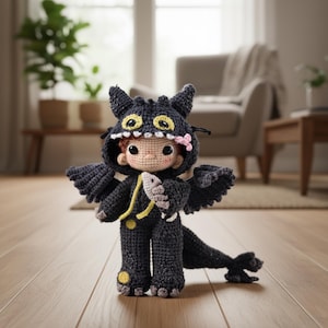 May include: A crocheted doll dressed as a dragon, featuring a black hooded jumpsuit with yellow accents, wings, and a tail. The doll has a light skin tone, rosy cheeks, and a pink flower. It holds a fish.