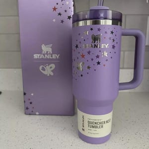 Peut inclure: Un gobelet Stanley Quencher H2.0 lavande avec une poign&eacute;e, un couvercle et une paille, &agrave; c&ocirc;t&eacute; d'une bo&icirc;te assortie. Le gobelet et la bo&icirc;te sont d&eacute;cor&eacute;s d'&eacute;toiles argent&eacute;es. Le gobelet porte le logo Stanley et le texte "QUENCHER H2.0 TUMBLER".