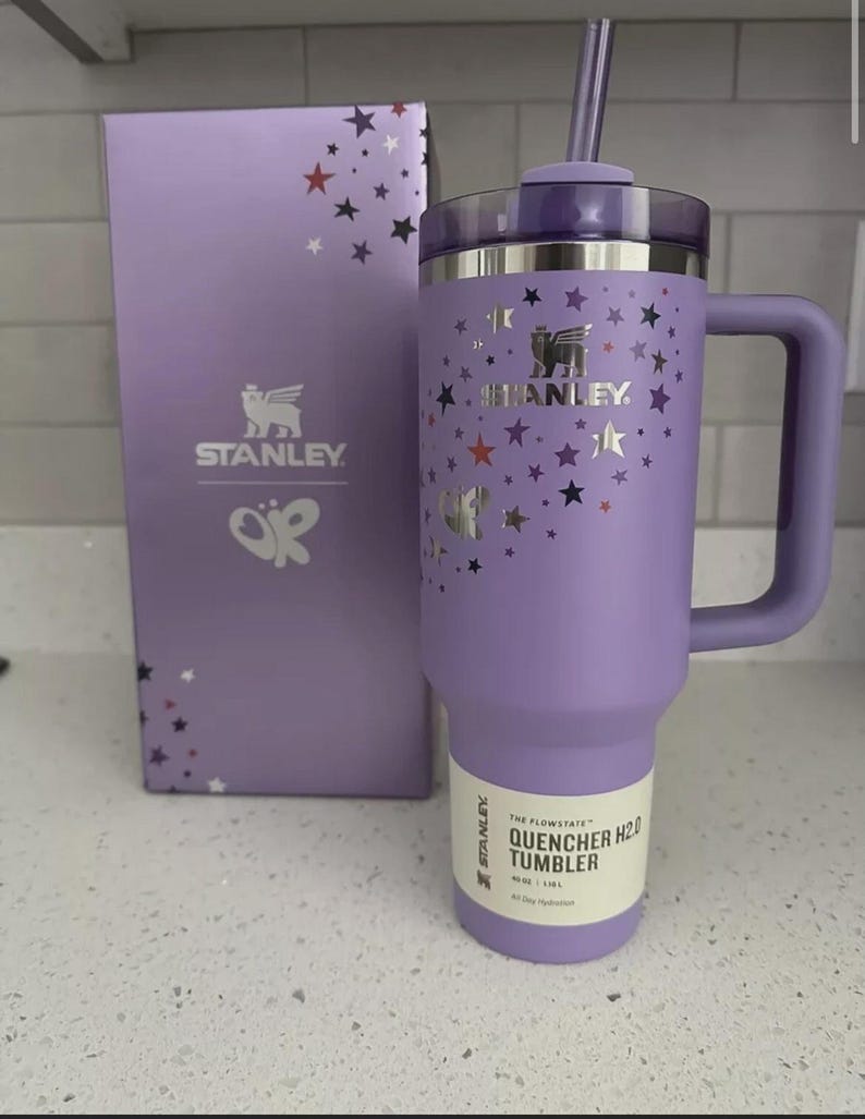 Peut inclure: Un gobelet Stanley Quencher H2.0 lavande avec une poign&eacute;e, un couvercle et une paille, &agrave; c&ocirc;t&eacute; d'une bo&icirc;te assortie. Le gobelet et la bo&icirc;te sont d&eacute;cor&eacute;s d'&eacute;toiles argent&eacute;es. Le gobelet porte le logo Stanley et le texte "QUENCHER H2.0 TUMBLER".