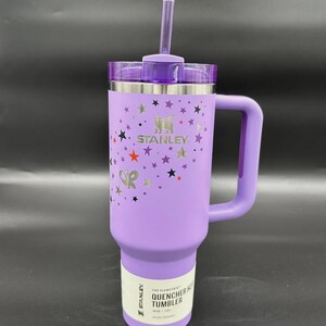 Peut inclure: Un gobelet Stanley Quencher H2.0 Flowstate violet avec une poign&eacute;e et une paille. Le gobelet pr&eacute;sente un motif d'&eacute;toiles et le logo Stanley. L'&eacute;tiquette indique "Quencher H2.0 Tumbler".