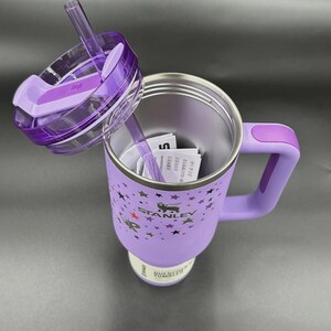 Peut inclure: Un gobelet isotherme violet avec une poign&eacute;e et un couvercle transparent. Le gobelet a une paille et est d&eacute;cor&eacute; d'&eacute;toiles et du logo Stanley. Le couvercle est partiellement ouvert.