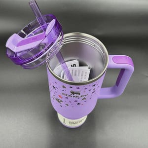 Peut inclure: Un gobelet isotherme violet avec une poign&eacute;e et un couvercle transparent avec une paille. Le gobelet a un corps violet clair avec un motif d'&eacute;toiles et le logo Stanley. Le couvercle est ouvert.