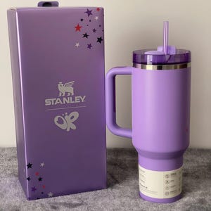 Peut inclure: Un gobelet Stanley lavande avec une poign&eacute;e, un couvercle et une paille, &agrave; c&ocirc;t&eacute; d'une bo&icirc;te assortie. La bo&icirc;te pr&eacute;sente le logo Stanley, des &eacute;toiles et les lettres "OR". Le gobelet a une capacit&eacute; d'environ 1,2 litre.