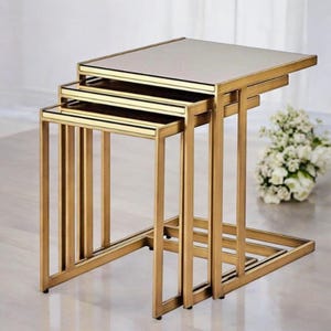 May include: A set of three nesting tables with a gold metal frame and mirrored tops. The tables are rectangular and vary in size, designed to fit one under the other. The design is modern and elegant.