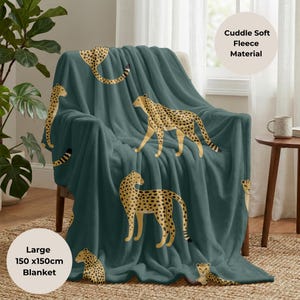 May include: A teal fleece blanket with a cheetah pattern draped over a chair. The blanket features multiple cheetah illustrations in tan and black. The blanket is labeled as a large 150 x 150cm blanket and made of cuddle soft fleece material.