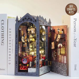 Könnte beinhalten: Ein Miniatur-DIY-Bastelset eines Dioramas eines Bahnhofs, mit einem roten Zug, Backsteinmauern und dekorativen Elementen. Das Set enthält den Text "DIY CRAFTS KITS" auf einem kreisförmigen Etikett. Das Diorama wird von Büchern mit den Titeln "CLOTHING" und "Person of the Year" flankiert.