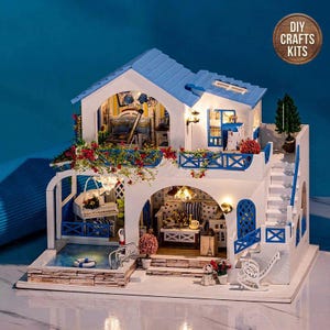 Puede incluir: Una casa de muñecas en miniatura de bricolaje con temática mediterránea. La estructura de dos pisos presenta un techo azul, paredes blancas y detalles azules. Incluye una pequeña piscina, balcón y mobiliario interior detallado. La imagen incluye el texto "DIY Crafts Kits".