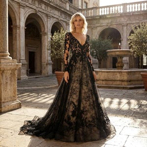 Op de afbeelding: Een zwarte kanten trouwjurk met lange mouwen en een diepe V-hals. De jurk heeft gedetailleerde bloemenkant over een nude voering, wat een opvallend contrast geeft. De jurk heeft een zwierige rok en een lange sleep, geschikt voor een formele gelegenheid.