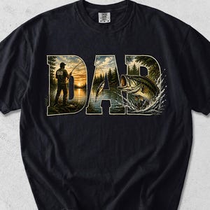 May include: Black t-shirt with a graphic design featuring the word "DAD" in a stylized font. The letters are filled with a fishing scene, including a person fishing at sunset and a leaping fish. The shirt is made of a soft material.