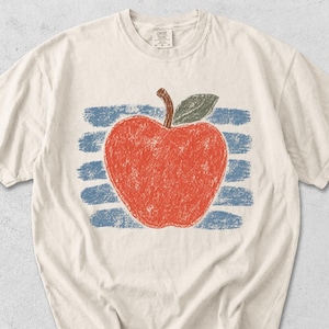 Puede incluir: Camiseta color crema con una ilustración de una manzana roja, hoja verde y tallo marrón. La manzana está sobre un fondo de rayas horizontales azules. El diseño tiene un estilo de dibujo a mano. La camiseta tiene mangas cortas y un corte holgado.